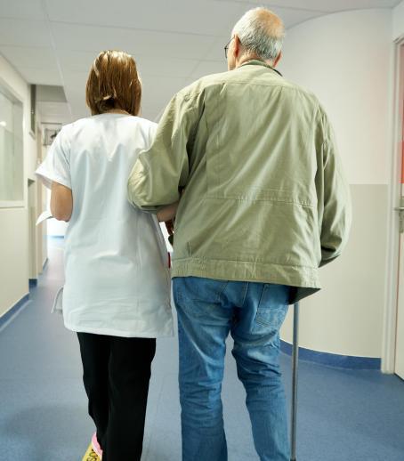 Une soignante marche de dos dans un couloir en tenant le bras d'un patient