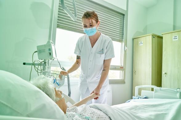 Une soignante et une patiente se donnent la main dans une chambre alors que la patiente est allongée dans un lit - Agrandir l'image, fenêtre modale