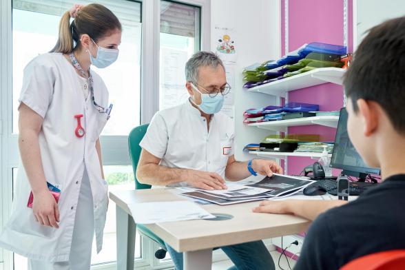 Un docteur et une soignante regardent la radio d'un jeune patient présent avec eux dans un bureau - Agrandir l'image, fenêtre modale